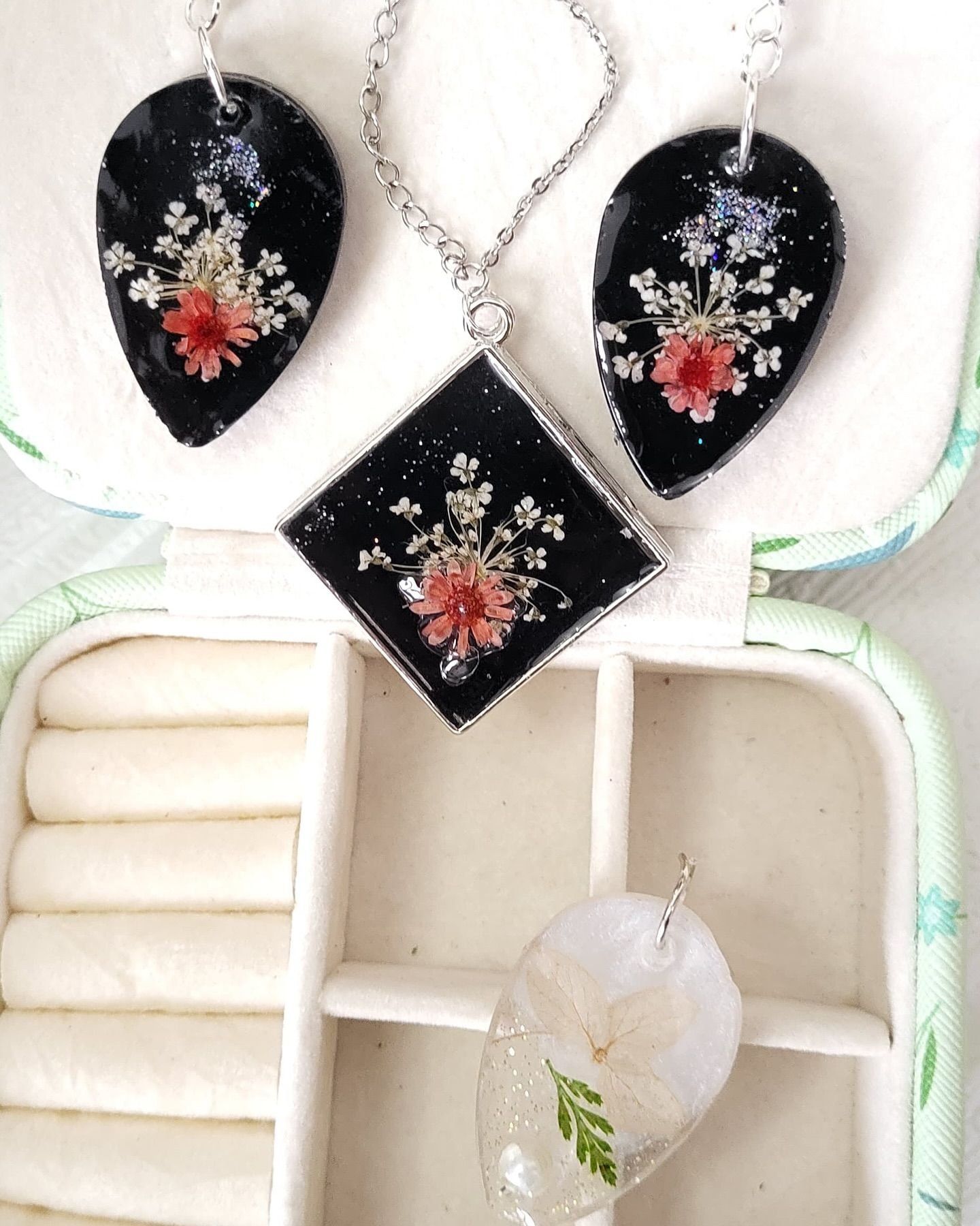 Two black teardrops and a square pendant, all set with white Queen Anne’s lace, one coral daisy and a scatter of silver glitter, laid on the cream lining of a jewellery case with their silver chains coiled between them