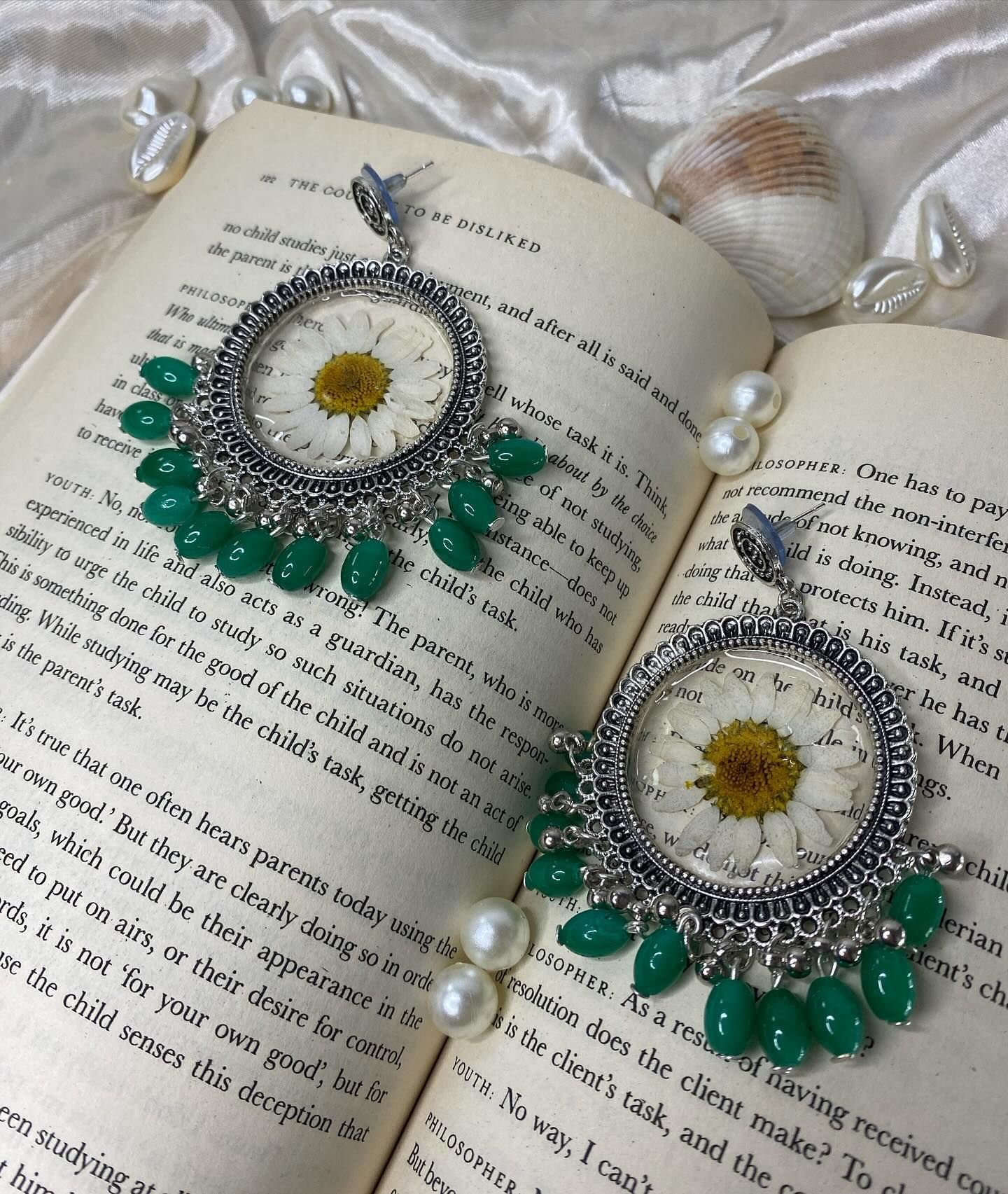 Two silver jhumkas, each set with a pressed white daisy under clear resin and fringed with green beads, lying on the open pages of a book with freshwater pearls and a seashell