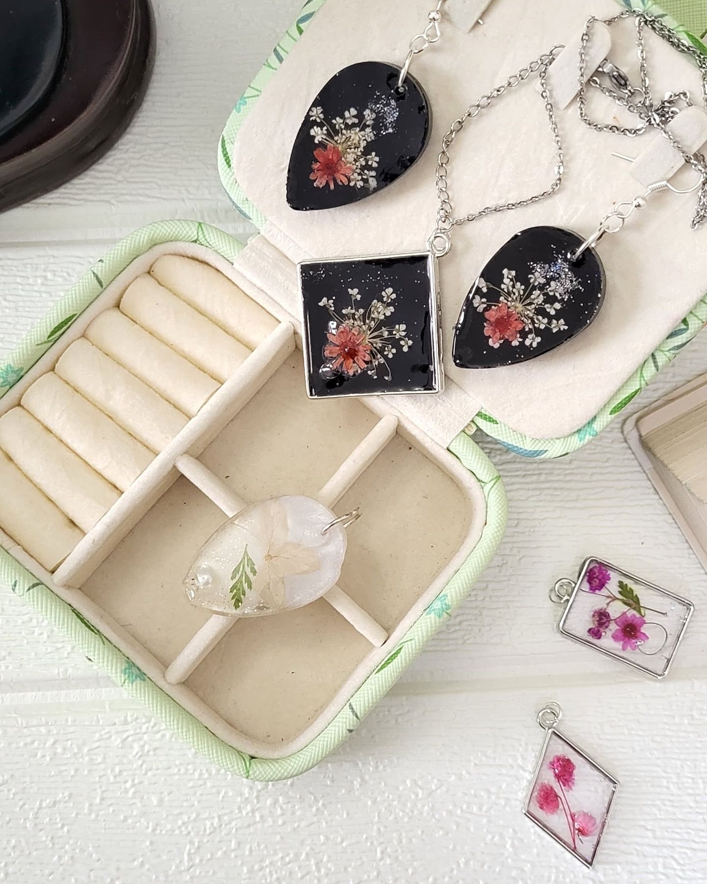 A group of resin pendants and earrings arranged in and around an open jewellery box: black teardrop drops set with a coral flower and white baby’s breath, a square black pendant on a silver chain, a pale oval pendant holding a fern frond, and two small clear pendants set with pink and magenta blooms