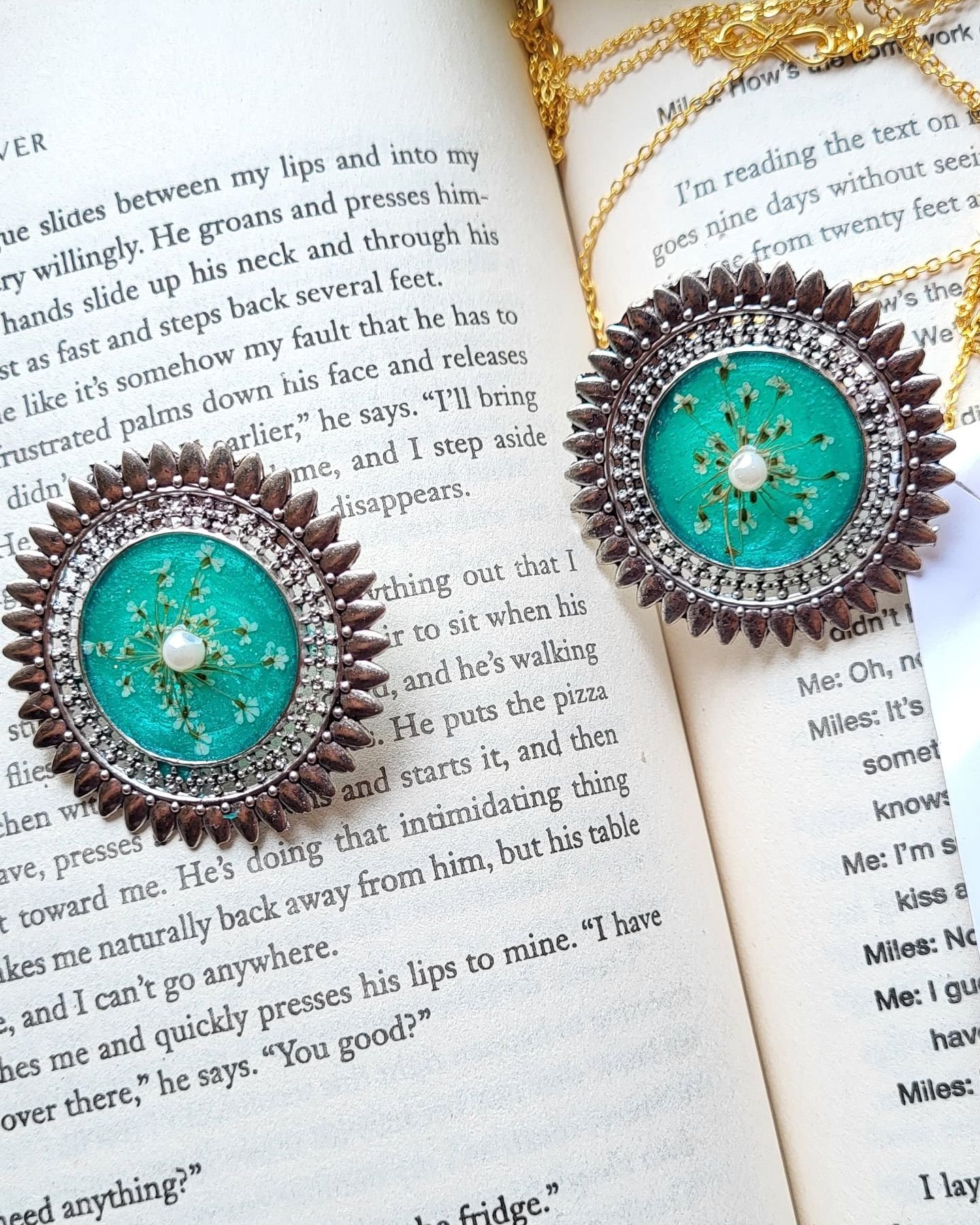 A pair of round earrings set with teal resin, a spray of pressed white baby’s breath and a single pearl at the centre, framed in ornate oxidised silver and dark bronze, lying on the open pages of a book with fine gold chains